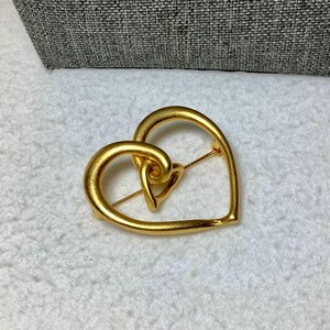 May include: A gold-toned heart-shaped brooch with a smaller heart design inside. The brooch is made of metal and has a brushed finish.