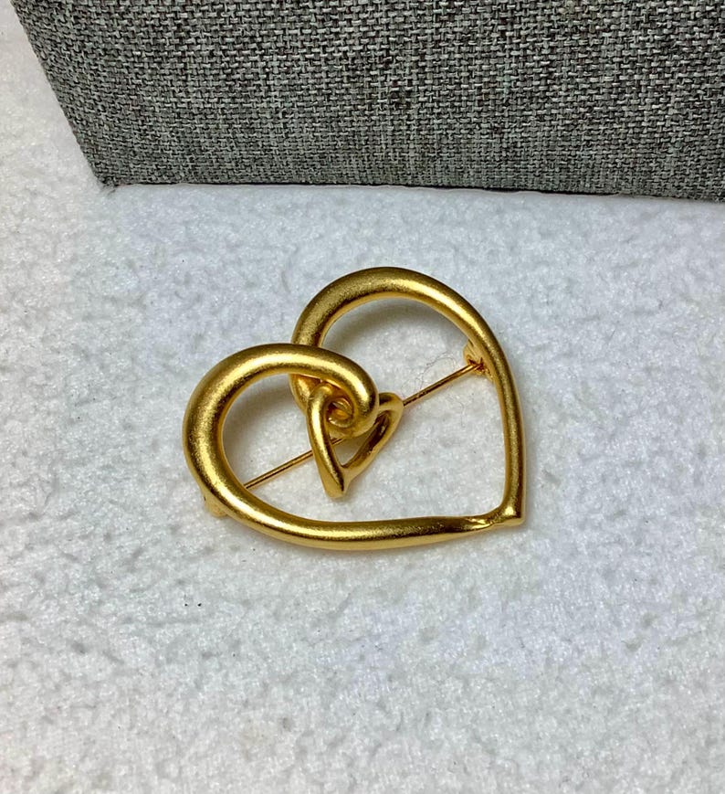 May include: A gold-toned heart-shaped brooch with a smaller heart design inside. The brooch is made of metal and has a brushed finish.