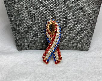 May include: A red, white and blue ribbon pin with rhinestones. The pin is gold-toned and has a safety clasp.