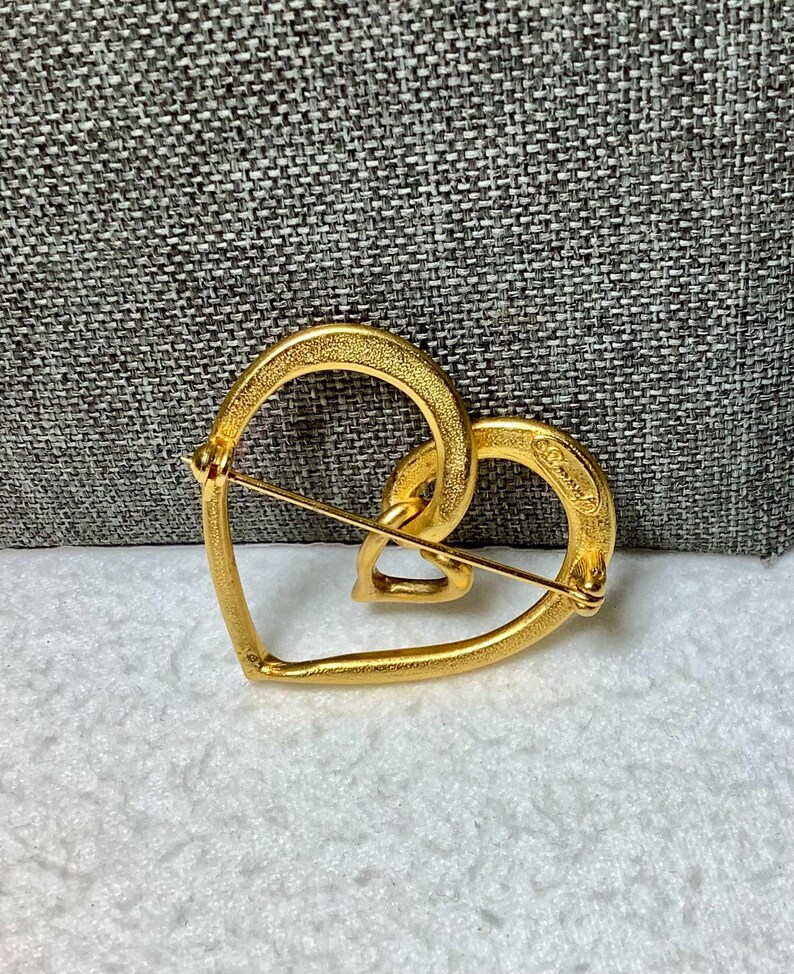 May include: Gold-toned heart-shaped brooch with a smaller heart design inside. The brooch is marked "Coro" on the back.