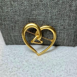May include: A gold-toned heart-shaped brooch with an intricate design. The heart is made of two intertwined loops.