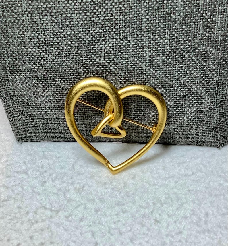 May include: A gold-toned heart-shaped brooch with an intricate design. The heart is made of two intertwined loops.