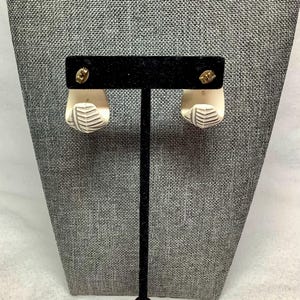May include: A pair of off-white, geometric earrings with a leaf-like design. The earrings are displayed on a black stand against a gray textured background. The earrings have gold-colored posts.