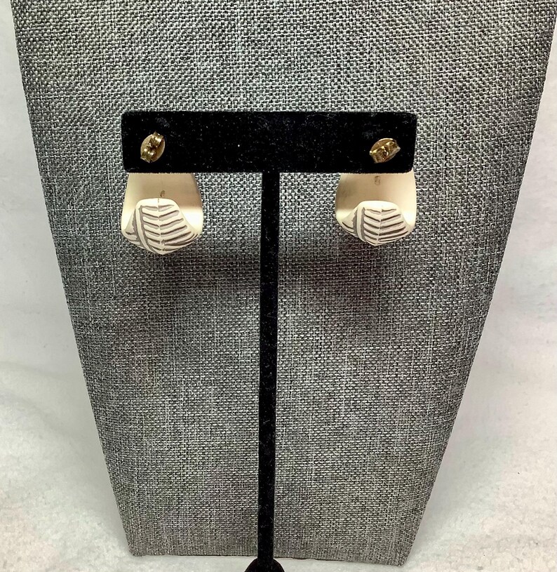 May include: A pair of off-white, geometric earrings with a leaf-like design. The earrings are displayed on a black stand against a gray textured background. The earrings have gold-colored posts.