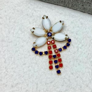 May include: A gold-tone brooch with a white flower design and a red, white, and blue tassel. The flower has five petals and a center stone.
