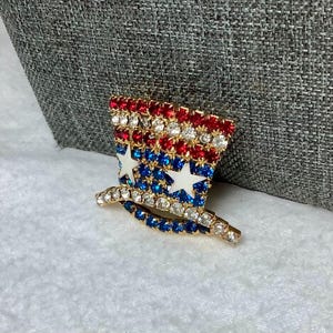 May include: A gold-tone pin shaped like a top hat with red, white, and blue rhinestones arranged in a pattern resembling the American flag.
