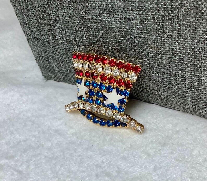 May include: A gold-tone pin shaped like a top hat with red, white, and blue rhinestones arranged in a pattern resembling the American flag.