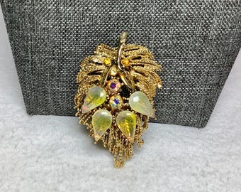 Könnte beinhalten: Eine goldfarbene Brosche mit einem Blattdesign und schillernden Glassteinen. Die Brosche ist mit kleineren goldenen Perlen verziert und hat eine herabhängende Kette.