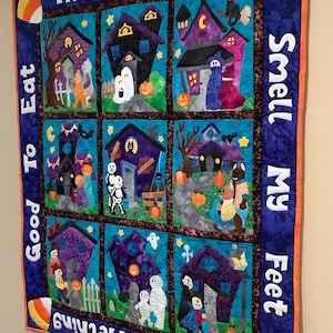K&ouml;nnte beinhalten: Ein Halloween-Quilt mit neun quadratischen Feldern, die Spukh&auml;user, Geister und Trick-or-Treat-Kinder darstellen. Der Quilt zeigt die S&auml;tze "Trick or Treat", "Good To Eat" und "Give Me Something Smell My Feet" in wei&szlig;er Schrift auf blauem Hintergrund.