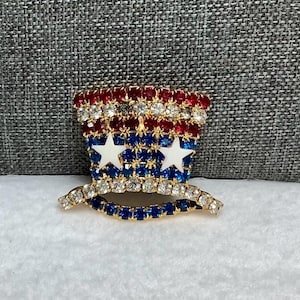May include: A red, white, and blue rhinestone brooch in the shape of a top hat. The hat has two white stars on the front.