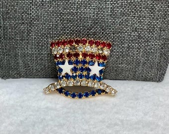May include: A red, white and blue rhinestone brooch in the shape of a top hat. The hat has two white stars on the front.