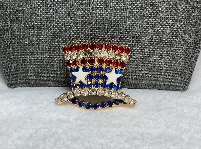 May include: A red, white, and blue rhinestone brooch in the shape of a top hat. The hat has two white stars on the front.