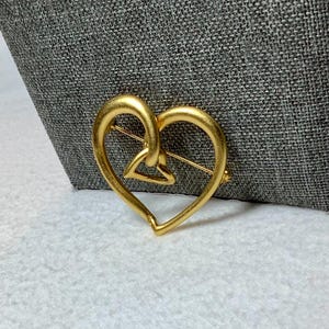 May include: A gold-toned heart-shaped brooch with a smaller heart design inside. The brooch is made of metal and has a pin on the back.