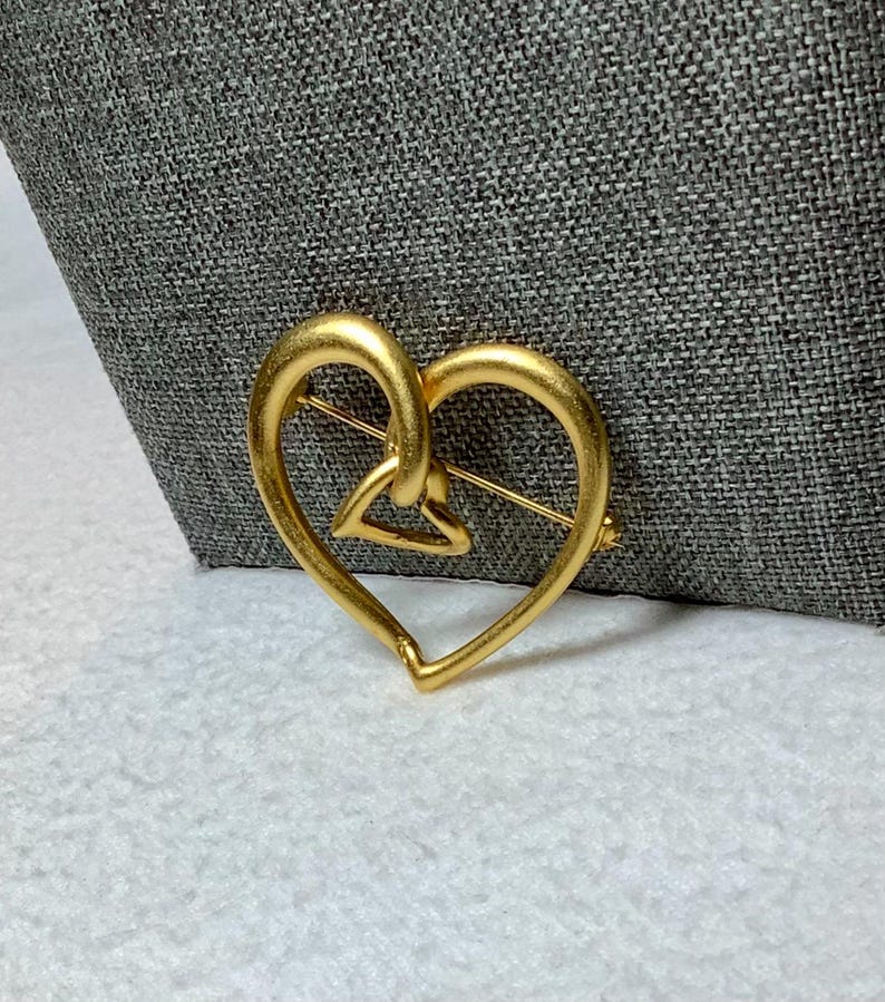 May include: A gold-toned heart-shaped brooch with a smaller heart design inside. The brooch is made of metal and has a pin on the back.