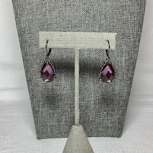 May include: A pair of drop earrings with a dark metal finish. Each earring features a faceted, teardrop-shaped purple gemstone. The gemstones are surrounded by a decorative metal border. The earrings are displayed on a gray jewelry stand.