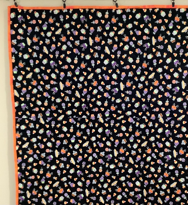 K&ouml;nnte beinhalten: Eine schwarze Steppdecke mit einem sich wiederholenden Muster von Halloween-S&uuml;&szlig;igkeiten, Cupcakes und Lollis in Orange, Lila und Wei&szlig;. Die Steppdecke hat einen orangefarbenen Rand und h&auml;ngt an Gardinenringen.