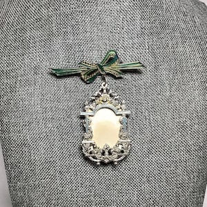 May include: A silver-toned brooch with a white cameo in a decorative frame. The brooch is accented with green and gold enamel and features a green and gold bow.