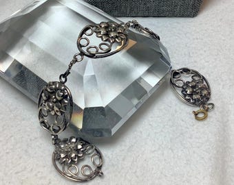 May include: A silver bracelet with a floral design. The bracelet has three links, each with a different floral pattern. The bracelet is on a white surface.
