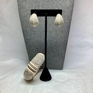 May include: A cream-colored jewelry set featuring earrings and a bracelet. The earrings are teardrop-shaped with a geometric pattern in black and white. The bracelet has a similar design with cream, black, and brown stripes. The jewelry is displayed on a black stand.