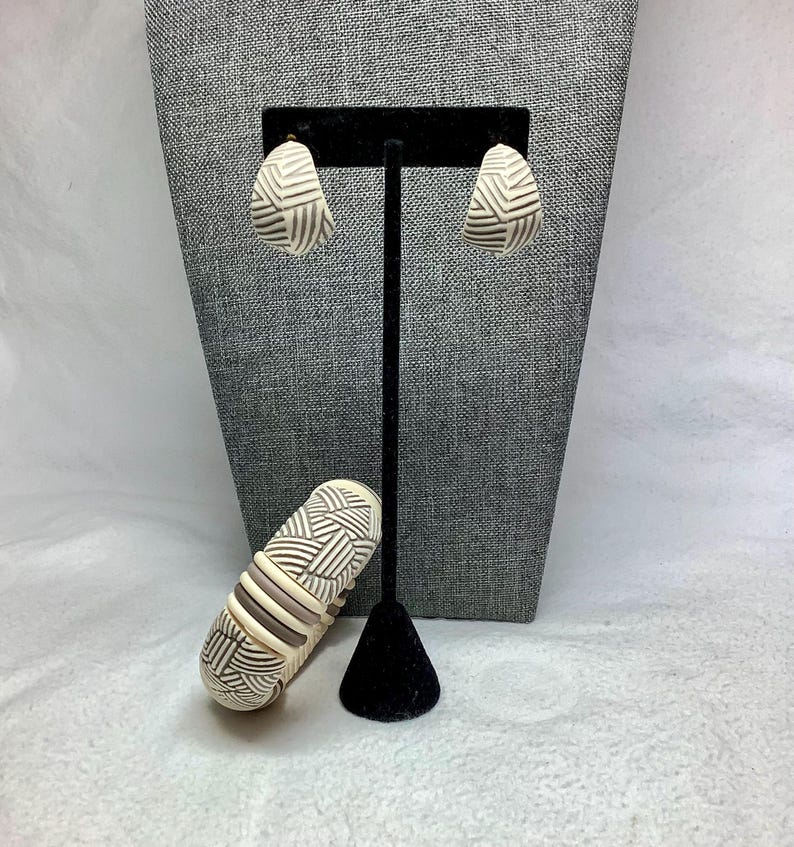 May include: A cream-colored jewelry set featuring earrings and a bracelet. The earrings are teardrop-shaped with a geometric pattern in black and white. The bracelet has a similar design with cream, black, and brown stripes. The jewelry is displayed on a black stand.