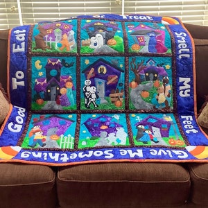 K&ouml;nnte beinhalten: Ein Halloween-Quilt mit neun quadratischen Feldern, die Spukh&auml;user, Trick-or-Treat-Kinder und K&uuml;rbisse zeigen. Der Quilt hat einen blauen Rand mit den Phrasen "Good To Eat", "Give Me Something" und "Smell My Feet". Der Quilt liegt auf einem braunen Sofa.