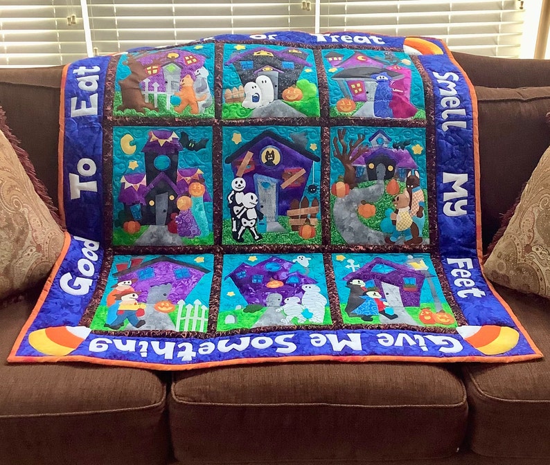 K&ouml;nnte beinhalten: Ein Halloween-Quilt mit neun quadratischen Feldern, die Spukh&auml;user, Trick-or-Treat-Kinder und K&uuml;rbisse zeigen. Der Quilt hat einen blauen Rand mit den Phrasen "Good To Eat", "Give Me Something" und "Smell My Feet". Der Quilt liegt auf einem braunen Sofa.