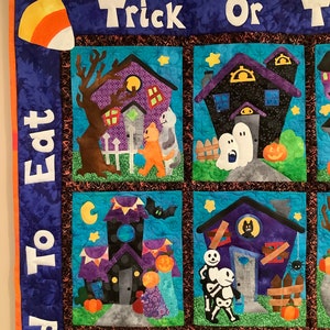 K&ouml;nnte beinhalten: Ein Halloween-Quilt mit blauem Hintergrund und orangefarbener Umrandung. Der Quilt zeigt mehrere Quadrate mit Bildern von Spukh&auml;usern, Geistern, K&uuml;rbissen und Trick-or-Treatern. Die Worte "Trick or Treat" und "Eat To Good" sind zu sehen.