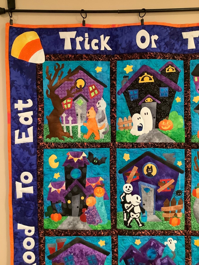 K&ouml;nnte beinhalten: Ein Halloween-Quilt mit blauem Hintergrund und orangefarbener Umrandung. Der Quilt zeigt mehrere Quadrate mit Bildern von Spukh&auml;usern, Geistern, K&uuml;rbissen und Trick-or-Treatern. Die Worte "Trick or Treat" und "Eat To Good" sind zu sehen.