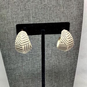 May include: A pair of cream-colored, half-hoop earrings with a geometric, striped design in dark brown. The earrings are displayed on a black jewelry stand against a gray textured background. The earrings have a post-back closure.