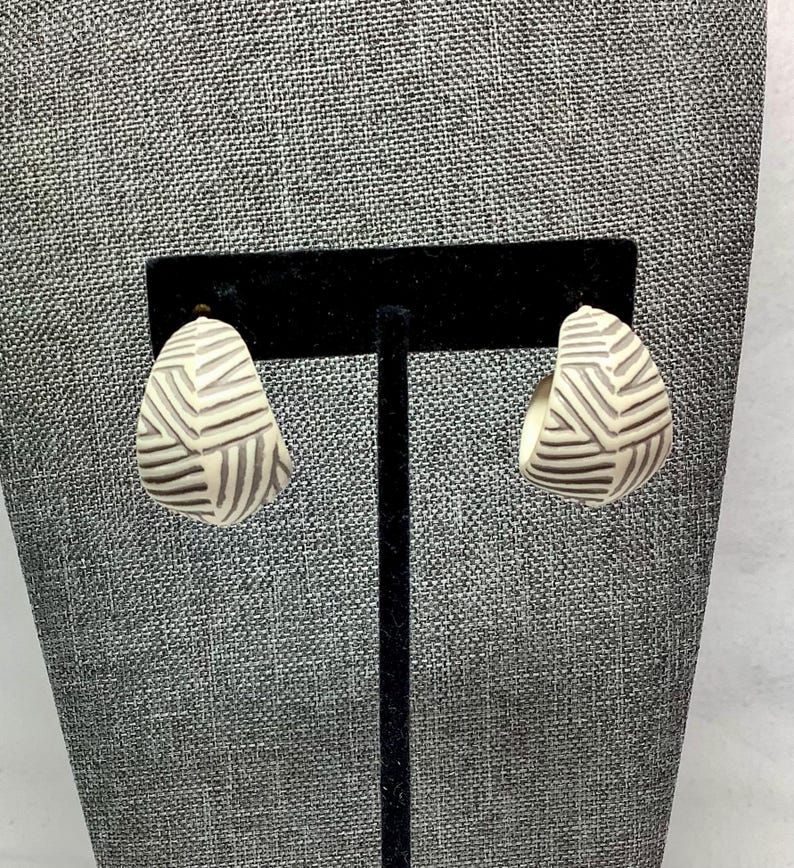 May include: A pair of cream-colored, half-hoop earrings with a geometric, striped design in dark brown. The earrings are displayed on a black jewelry stand against a gray textured background. The earrings have a post-back closure.
