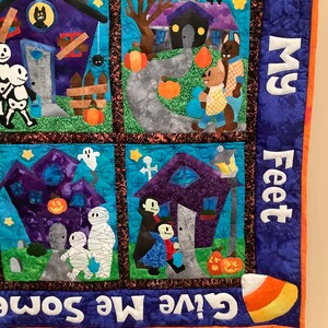 K&ouml;nnte beinhalten: Ein Halloween-Patchwork-Quilt mit vier quadratischen Feldern, die H&auml;user, Skelette, Mumien und Vampire zeigen. Der Rand enth&auml;lt den Text "Give Me Some..." und "...ll My Feet" sowie eine Grafik von einem Candy Corn.