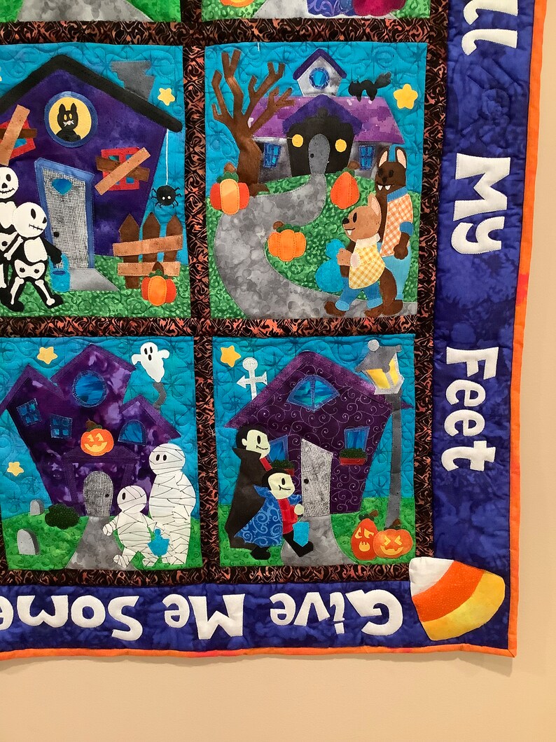 K&ouml;nnte beinhalten: Ein Halloween-Patchwork-Quilt mit vier quadratischen Feldern, die H&auml;user, Skelette, Mumien und Vampire zeigen. Der Rand enth&auml;lt den Text "Give Me Some..." und "...ll My Feet" sowie eine Grafik von einem Candy Corn.