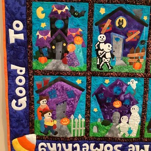 K&ouml;nnte beinhalten: Ein Stoffquilt zum Thema Halloween mit mehreren quadratischen Feldern, die applizierte Szenen von Geistern, Skeletten, Mumien und K&uuml;rbissen zeigen. Der blaue Rand zeigt den wei&szlig;en Text "Good To" und "Me Something".
