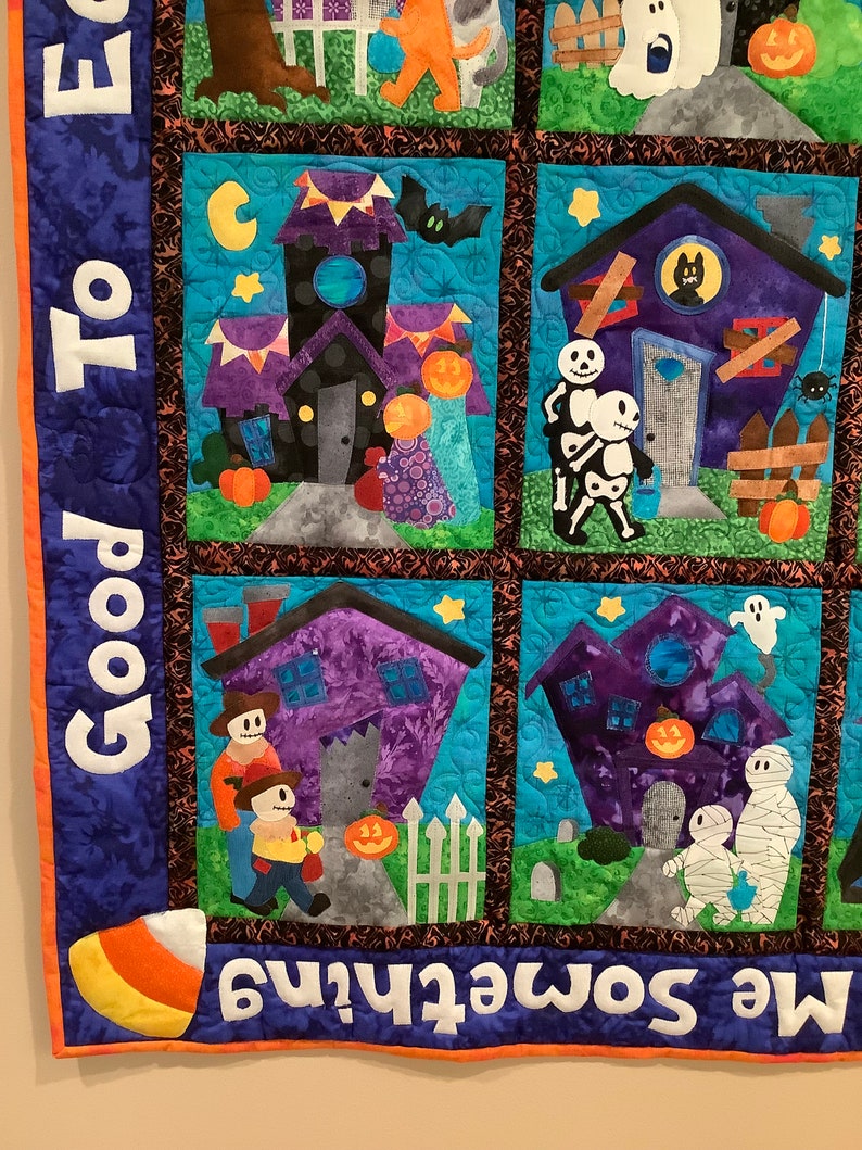 K&ouml;nnte beinhalten: Ein Stoffquilt zum Thema Halloween mit mehreren quadratischen Feldern, die applizierte Szenen von Geistern, Skeletten, Mumien und K&uuml;rbissen zeigen. Der blaue Rand zeigt den wei&szlig;en Text "Good To" und "Me Something".