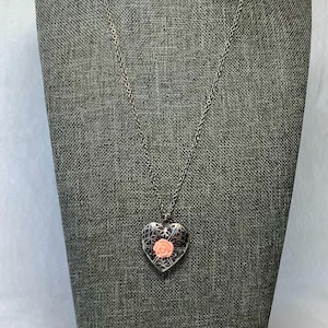 May include: A silver-toned heart-shaped locket necklace with a delicate filigree design. The locket features a pink rose in the center. The necklace is displayed on a gray mannequin.
