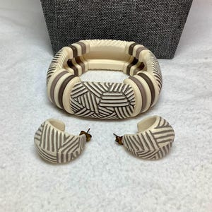 May include: A cream-colored bracelet and matching hoop earrings set. The bracelet features alternating cream and brown segments with a geometric design. The earrings have a similar pattern. The jewelry set is displayed on a white surface.