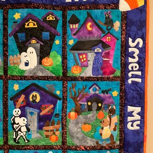 K&ouml;nnte beinhalten: Ein Halloween-Quilt mit mehreren quadratischen Feldern. Jedes Feld zeigt ein Spukhaus mit Geistern, K&uuml;rbissen und anderen gruseligen Figuren. Der Quilt tr&auml;gt die Aufschrift "Trick or Treat" und "Smell My Feet".