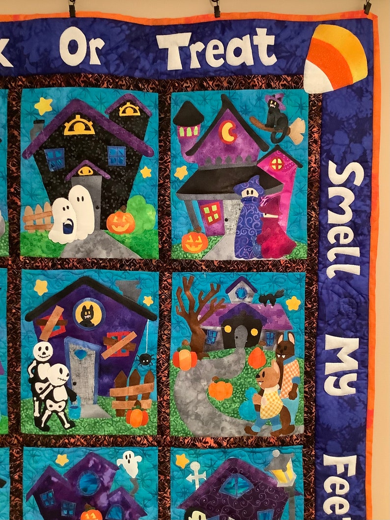 K&ouml;nnte beinhalten: Ein Halloween-Quilt mit mehreren quadratischen Feldern. Jedes Feld zeigt ein Spukhaus mit Geistern, K&uuml;rbissen und anderen gruseligen Figuren. Der Quilt tr&auml;gt die Aufschrift "Trick or Treat" und "Smell My Feet".