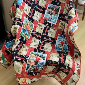 May include: A handmade quilt with a fishing theme. The quilt features a patchwork design with red, black, and beige squares. Some squares have vintage-style images of women fishing. The quilt is draped over a wooden chair.