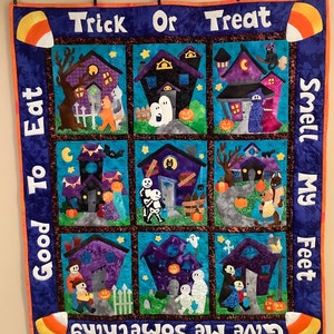 K&ouml;nnte beinhalten: Ein Halloween-Quilt mit neun quadratischen Feldern, die jeweils eine Spukhausszene zeigen. Der Quilt hat einen blauen Rand mit den Phrasen "Trick or Treat" und "Give Me Something to Eat Good Smell My Feet". Maiskolben-Akzente schm&uuml;cken die Ecken.
