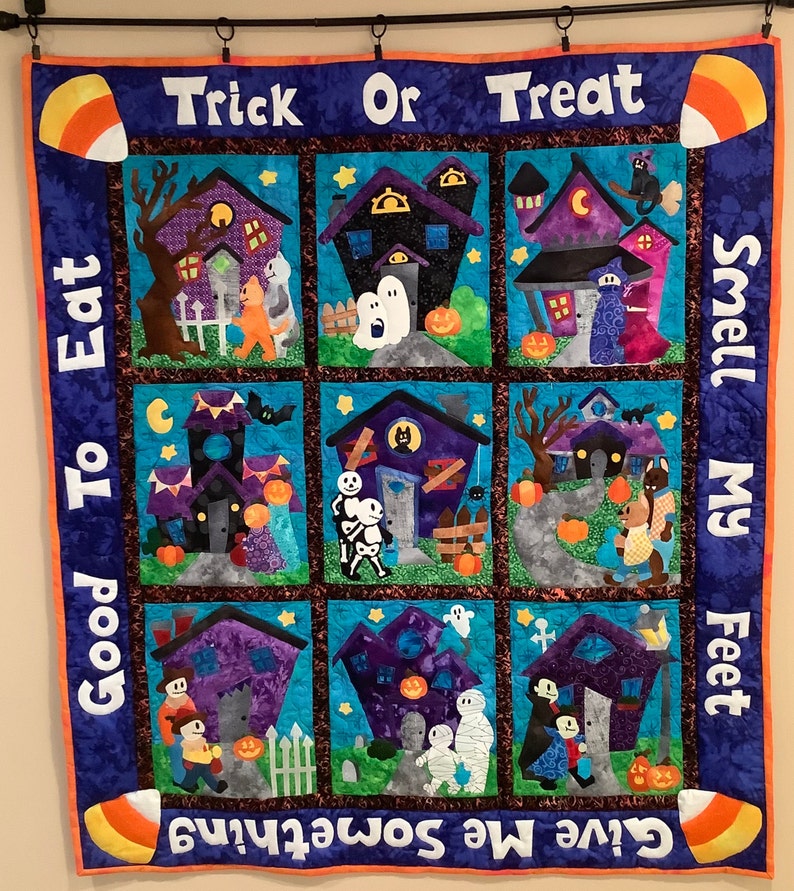 K&ouml;nnte beinhalten: Ein Halloween-Quilt mit neun quadratischen Feldern, die jeweils eine Spukhausszene zeigen. Der Quilt hat einen blauen Rand mit den Phrasen "Trick or Treat" und "Give Me Something to Eat Good Smell My Feet". Maiskolben-Akzente schm&uuml;cken die Ecken.