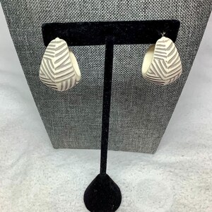 May include: A pair of cream-colored, teardrop-shaped earrings with a geometric gray pattern. The earrings are displayed on a black jewelry stand against a gray textured backdrop.