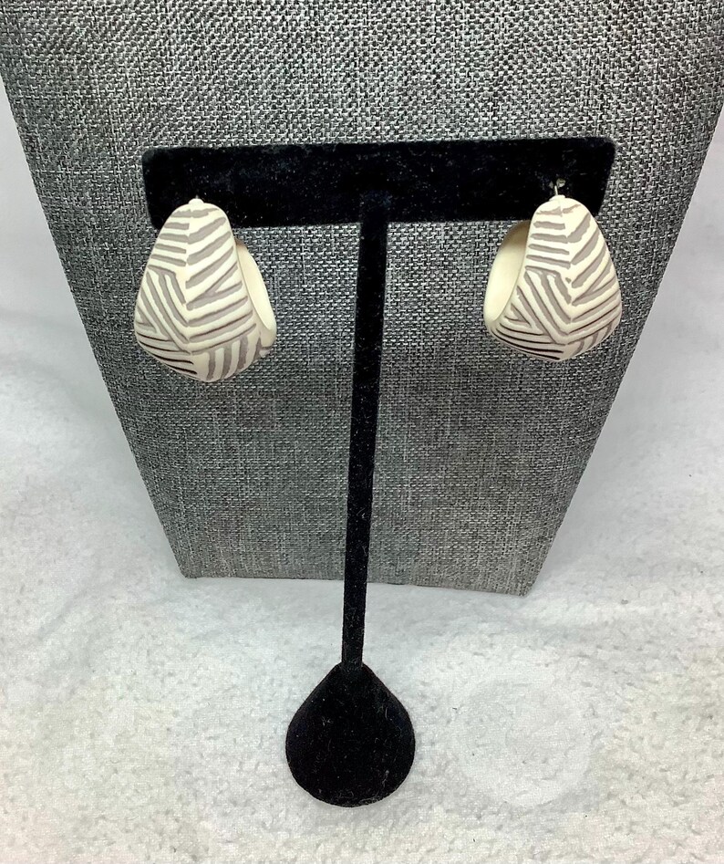 May include: A pair of cream-colored, teardrop-shaped earrings with a geometric gray pattern. The earrings are displayed on a black jewelry stand against a gray textured backdrop.