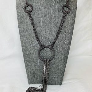 May include: A gunmetal-toned necklace with a large circular pendant and cascading fringe. The necklace features three smaller circular accents at the top and is displayed on a gray mannequin. The fringe is adorned with small, sparkling accents.