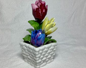 Könnte beinhalten: Ein weißer Keramik-Blumentopf mit geflochtener Textur, der drei Keramikblumen hält. Die Blumen sind rosa, gelb und blau.
