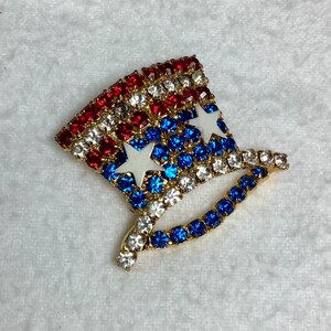 May include: A red, white, and blue jeweled pin shaped like a top hat with two white stars.
