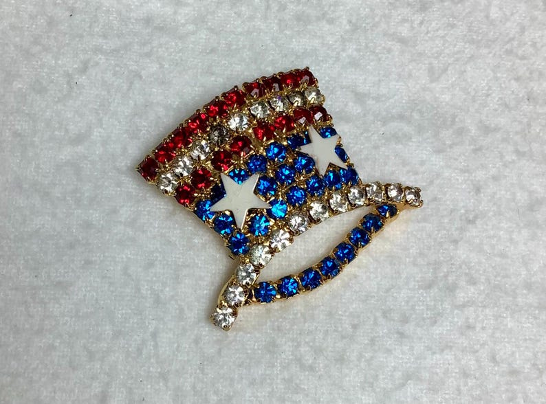 May include: A red, white, and blue jeweled pin shaped like a top hat with two white stars.