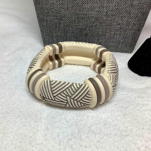 May include: Cream-colored bracelet with a geometric design. The bracelet features alternating sections of cream and gray-brown, with a textured pattern on the central rectangular pieces. The bracelet is made of a solid material and appears to be a stretch style.