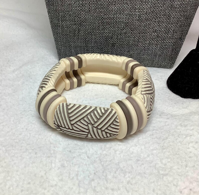 May include: Cream-colored bracelet with a geometric design. The bracelet features alternating sections of cream and gray-brown, with a textured pattern on the central rectangular pieces. The bracelet is made of a solid material and appears to be a stretch style.