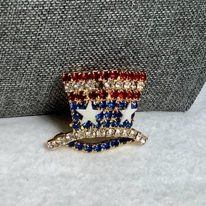 May include: A gold-toned pin with red, white, and blue rhinestones in the shape of a top hat with stars.